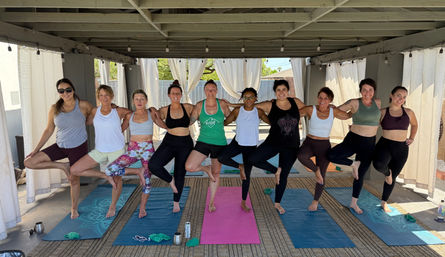 Eleven women smiling in tree pose during a sunny outdoor covered-patio yoga class on colorful mats, with sheer curtains and string lights overhead.