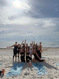 Group of women in black activewear cheering and posing on a sunny sandy beach with ocean backdrop, towels and water bottles in the foreground.