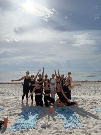 Group of women in black activewear cheering and posing on a sunny sandy beach with ocean backdrop, towels and water bottles in the foreground.