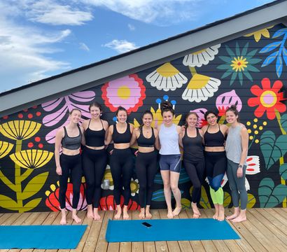 Eight women in yoga clothes standing barefoot on a rooftop wooden deck, smiling in front of a bright oversized floral mural with blue sky overhead and yoga mats on the floor.