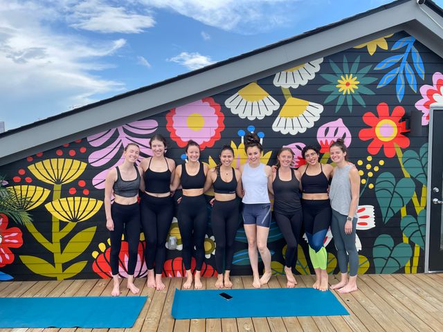 Eight women in yoga clothes standing barefoot on a rooftop wooden deck, smiling in front of a bright oversized floral mural with blue sky overhead and yoga mats on the floor.