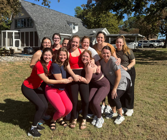 Group of smiling women in colorful activewear posing on a sunny suburban backyard lawn in front of a house