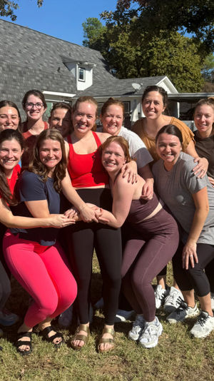 Group of smiling women in colorful activewear posing on a sunny suburban backyard lawn in front of a house