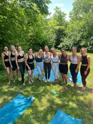 Outdoor yoga class in a sunny park: a group of women in athletic wear standing arm‑in‑arm on blue yoga mats surrounded by green trees and grass