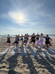 Sunlit beach yoga class: group of people doing tree pose on striped mats along the oceanfront at sunrise, calm sea and blue sky in the background.