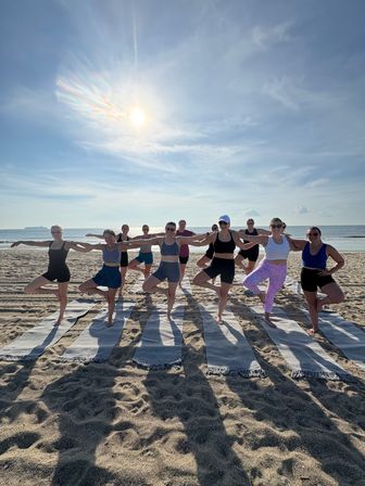Sunlit beach yoga class: group of people doing tree pose on striped mats along the oceanfront at sunrise, calm sea and blue sky in the background.