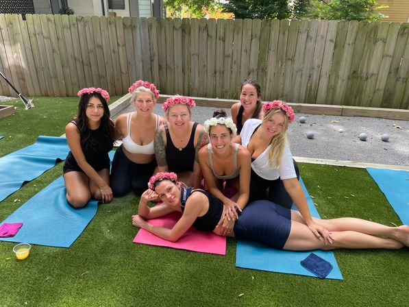 Seven women wearing flower crowns smiling and posing on colorful yoga mats on a fenced suburban backyard lawn during an outdoor yoga gathering.