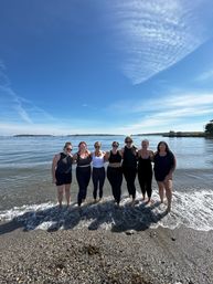 Eight friends standing arm-in-arm ankle-deep in gentle waves on a pebbly coastal beach, clear blue sky with wispy clouds and sailboats on the distant horizon.