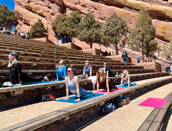 Outdoor yoga class on colorful mats spread across wooden benches in a red‑rock amphitheater with pine trees and bright blue sky in Colorado