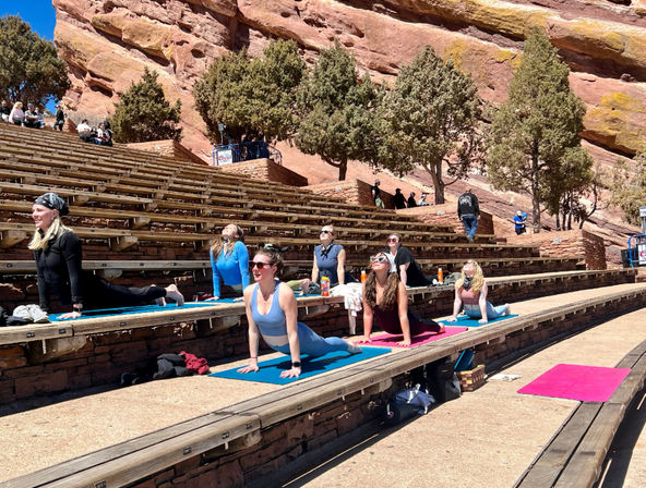 Outdoor yoga class on colorful mats spread across wooden benches in a red‑rock amphitheater with pine trees and bright blue sky in Colorado