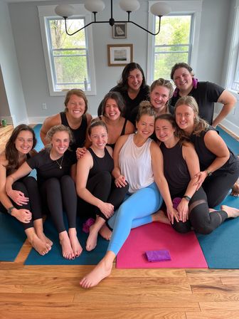 Smiling group of women in activewear gathered on colorful yoga mats in a bright home yoga studio with hardwood floors and large windows, post-class camaraderie.