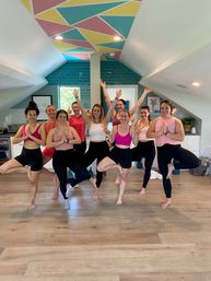 Smiling group yoga class of women practicing tree pose in a bright loft-style studio with hardwood floors, teal subway-tile accent wall and colorful geometric ceiling — indoor fitness/yoga session
