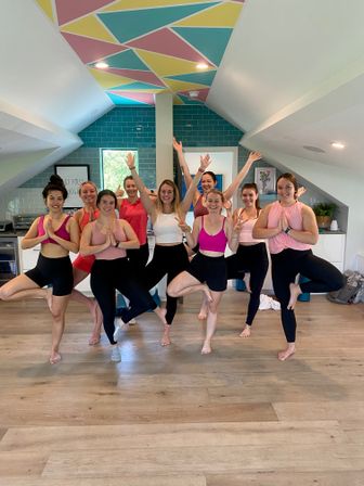 Smiling group yoga class of women practicing tree pose in a bright loft-style studio with hardwood floors, teal subway-tile accent wall and colorful geometric ceiling — indoor fitness/yoga session