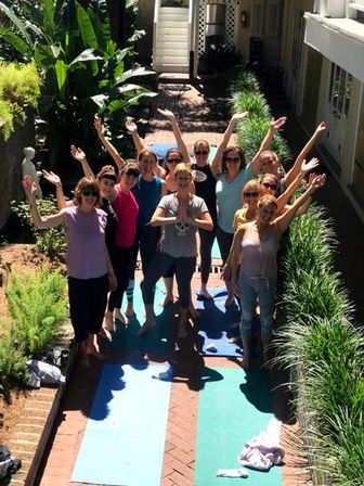 Outdoor courtyard yoga class: group of smiling women in casual activewear on colorful mats along a brick pathway, surrounded by lush greenery and raising their arms cheerfully.
