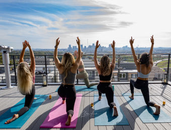 Energetic rooftop yoga session on a terrace with five people on mats in a lunge pose, arms raised toward a downtown city skyline under a blue morning sky.