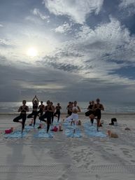 Group beach yoga class doing tree pose on blue towels at sunrise over the ocean, dramatic cloudy sky and calm waves on a sandy shore — peaceful oceanfront workout scene.