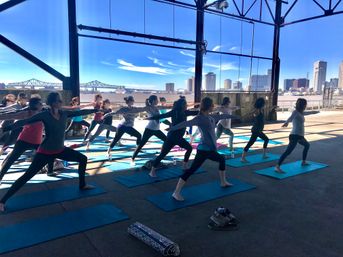Group outdoor yoga class doing warrior pose on blue mats in an open-air riverside pavilion with a bridge and city skyline across the river under a bright blue sky