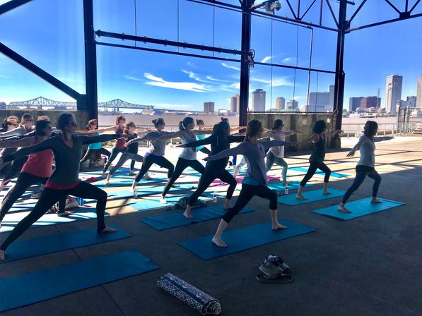 Group outdoor yoga class doing warrior pose on blue mats in an open-air riverside pavilion with a bridge and city skyline across the river under a bright blue sky