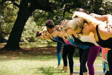 Row of women in colorful leggings and sunglasses leaning forward in a group outdoor yoga/workout class on a sunny park lawn