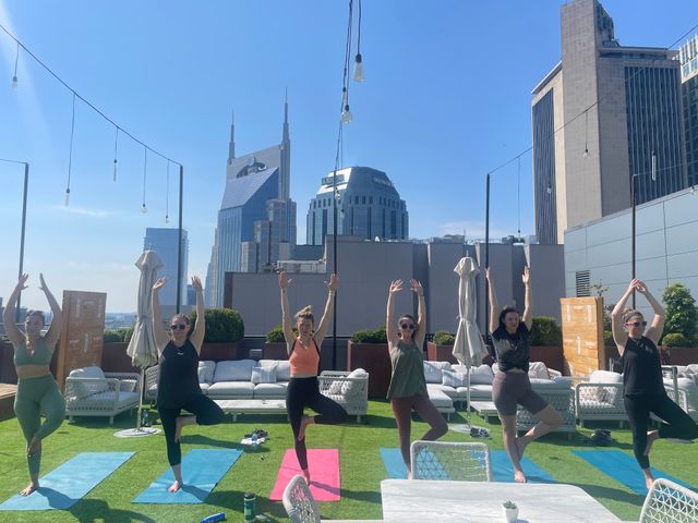 Rooftop yoga in Nashville: six people holding tree pose on colorful mats with downtown skyline and clear blue sky