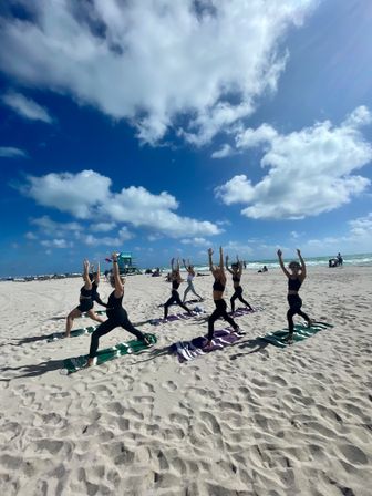 Group beach yoga class in black activewear on striped mats practicing Warrior pose on a sunlit sandy shore with turquoise ocean and a big blue sky dotted with fluffy clouds.
