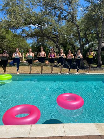 Group practicing yoga tree pose along a sunny backyard pool edge with turquoise water, pink and green inflatable rings, and oak trees under a blue sky.