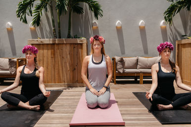 Three women wearing flower crowns meditate on yoga mats on a sunny wooden patio with tropical plants and lounge seating — outdoor group yoga and mindfulness session.