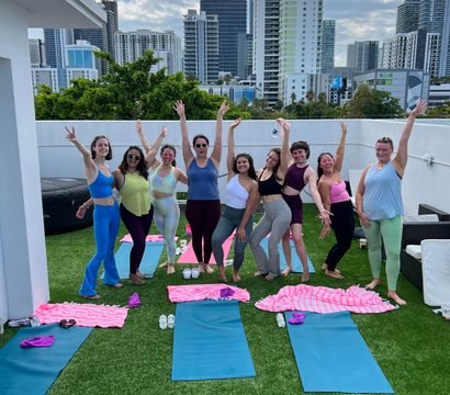 Nine women posing on colorful yoga mats during a rooftop yoga session on artificial turf, cheering with arms raised against an urban high-rise skyline and cloudy sky.