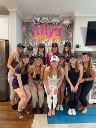 Smiling group of a dozen women in matching black caps posing in a living room for a bachelorette-style party in Nashville, in front of a handmade pink-and-gold sign and silver star balloons.
