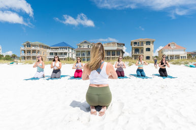 Sunny beach yoga class on white sand with an instructor kneeling in the foreground and six students on mats facing her, beachfront homes and blue sky in the background.