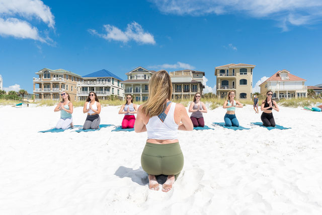 Sunny beach yoga class on white sand — instructor kneeling facing seven participants on mats with beachfront houses under a bright blue sky.