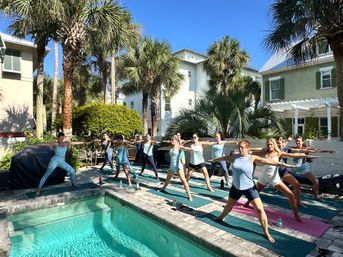 Sunlit poolside yoga class with a group striking warrior poses on mats by a turquoise pool, surrounded by palm trees and pastel coastal houses under a clear blue sky.