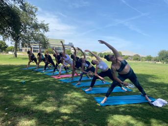 Group outdoor yoga class on blue mats performing side-angle stretches on a sunny green park lawn with trees and buildings in the background.