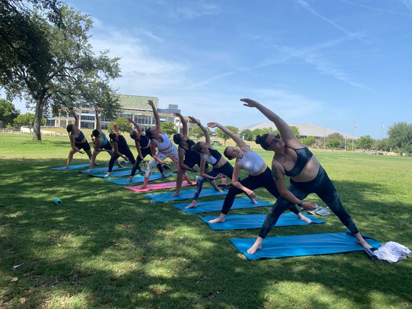 Group outdoor yoga class on blue mats performing side-angle stretches on a sunny green park lawn with trees and buildings in the background.