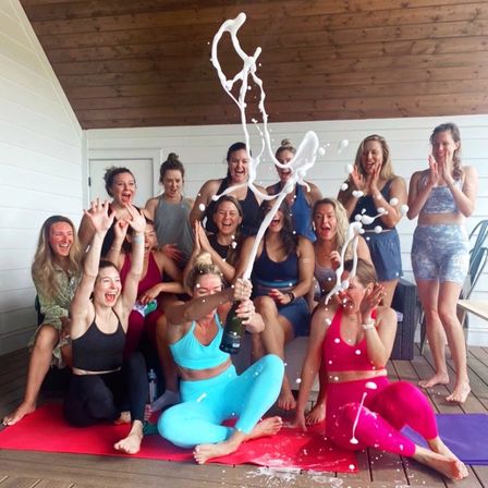 Happy group of women in colorful yoga outfits on a wooden deck celebrating together as champagne sprays into the air