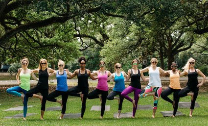 Outdoor yoga class with ten women standing arm-in-arm in tree pose on mats on a green lawn under large shade trees in a public park