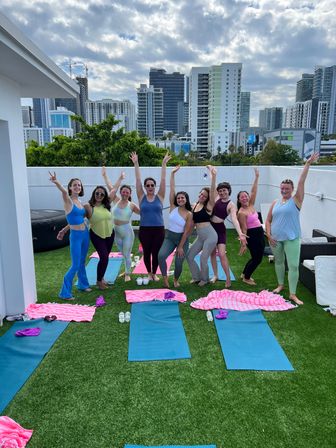 Rooftop yoga group: smiling women in colorful activewear strike joyful poses with arms raised on blue mats and pink towels on green turf, modern city skyline in the background