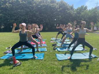 Sunlit outdoor yoga class in a park — a group of women on blue mats holding wide-legged poses on a green lawn with trees in the background.