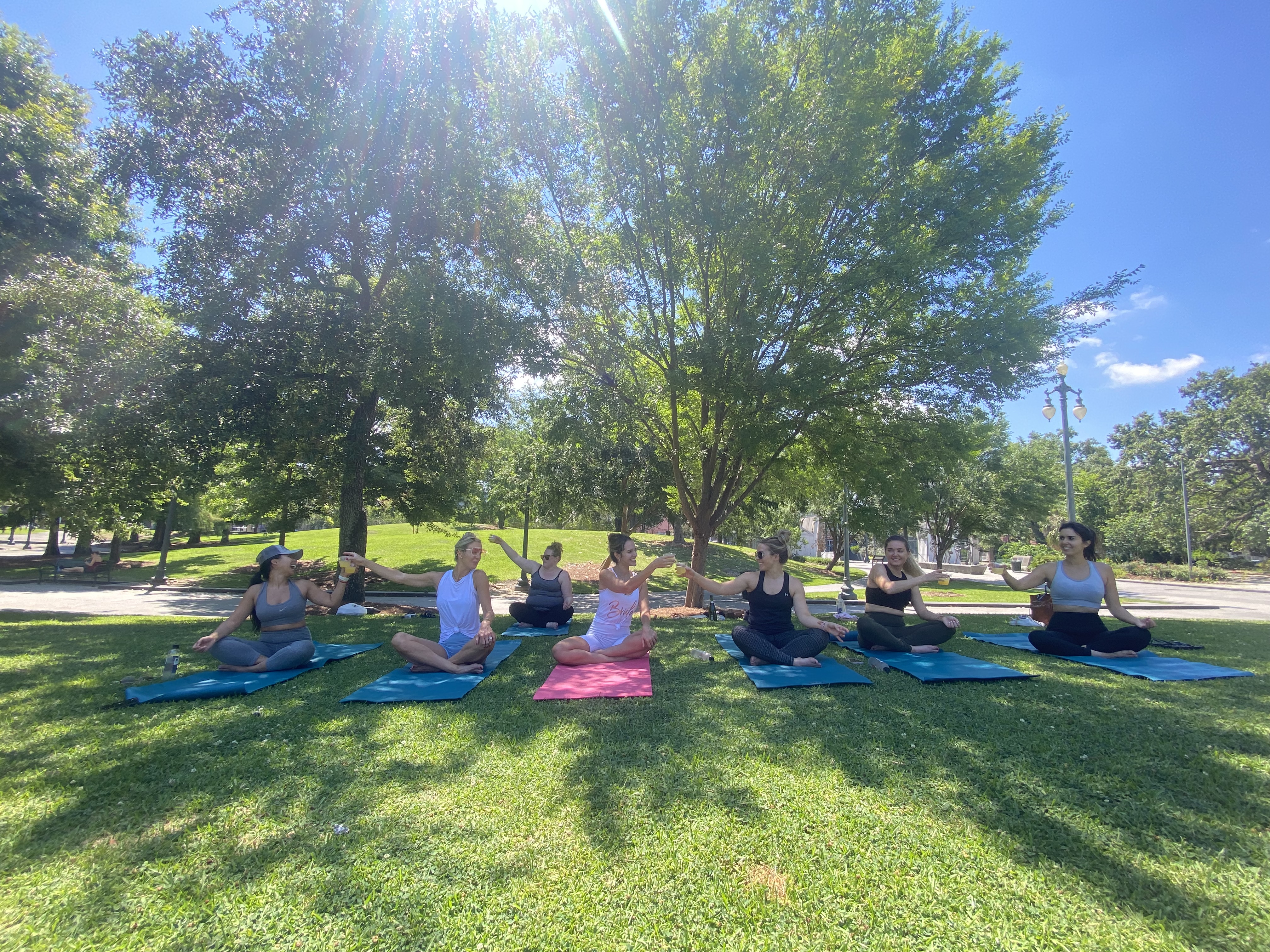 Seven women doing outdoor yoga on colorful mats in a sunny city park, seated and reaching hands under large leafy trees on a green lawn.