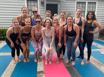 Group of women in colorful yoga gear smiling on blue and pink mats during an outdoor backyard yoga class on a white clapboard house patio.