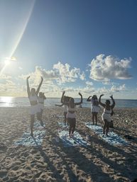 Sunlit beach workout: six women in matching activewear jumping with arms raised on towels by the ocean during golden hour under blue sky and fluffy clouds