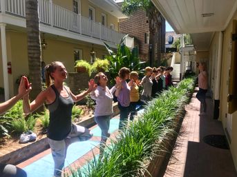 Sunny courtyard yoga class with a line of participants balancing in tree pose, palms pressed toward an instructor, surrounded by lush planters and a two-story balcony.