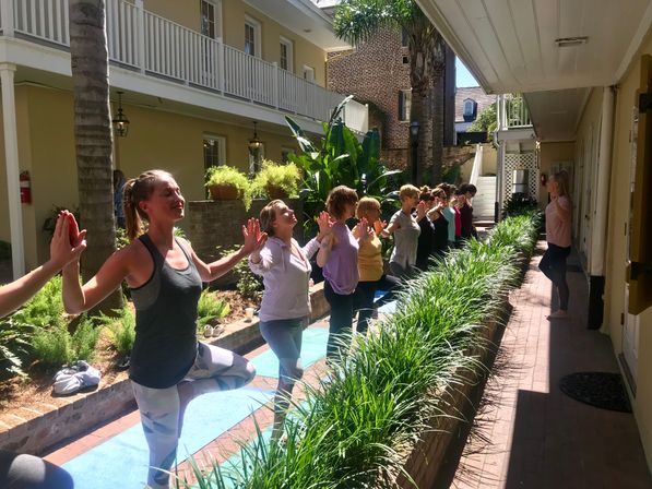 Sunny courtyard yoga class with a line of participants balancing in tree pose, palms pressed toward an instructor, surrounded by lush planters and a two-story balcony.