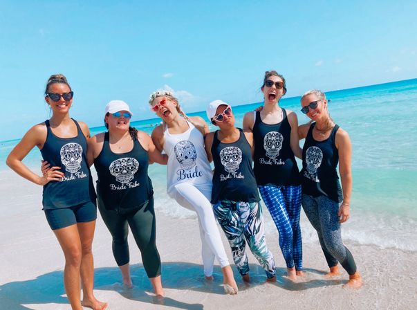 Cheerful bachelorette group of six women on a sunny turquoise beach, bride in white and friends in matching 'Bride Tribe' tank tops and sunglasses posing at the shoreline.