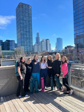 Seven smiling women with rolled yoga mats posing on a sunny rooftop deck with the Chicago skyline and Willis Tower visible behind modern high-rise buildings.