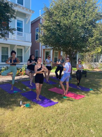 Group of women practicing outdoor yoga (tree pose) on colorful mats on a sunny residential lawn in front of pastel houses under a large shade tree