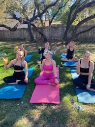 Group of six women seated on colorful yoga mats in a sunny backyard under large trees, smiling and holding mimosas after an outdoor yoga session and brunch-style celebration.