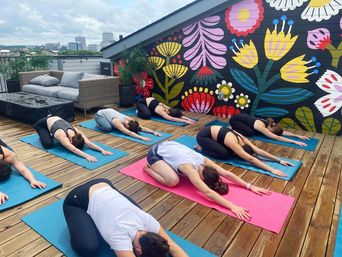Outdoor urban rooftop yoga class on a wooden deck, several people in child's pose on colorful mats in front of a vibrant floral mural, city skyline visible in the background.