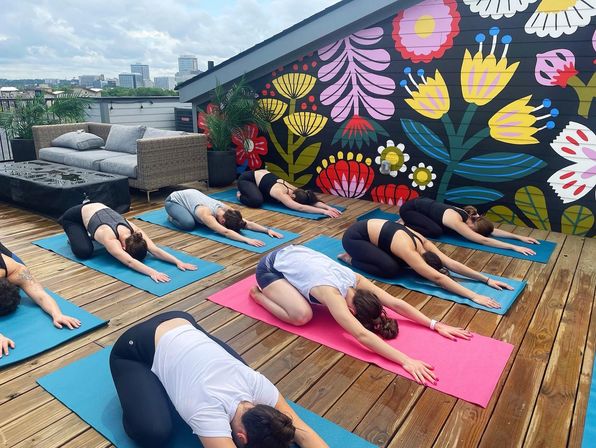 Outdoor urban rooftop yoga class on a wooden deck, several people in child's pose on colorful mats in front of a vibrant floral mural, city skyline visible in the background.