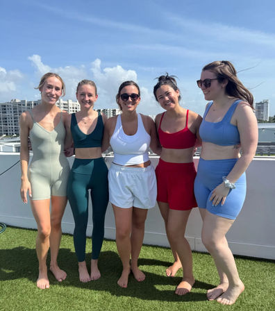 Five smiling friends in colorful active swimwear standing barefoot on a sunny rooftop terrace with coastal high-rise condos and blue sky in the background